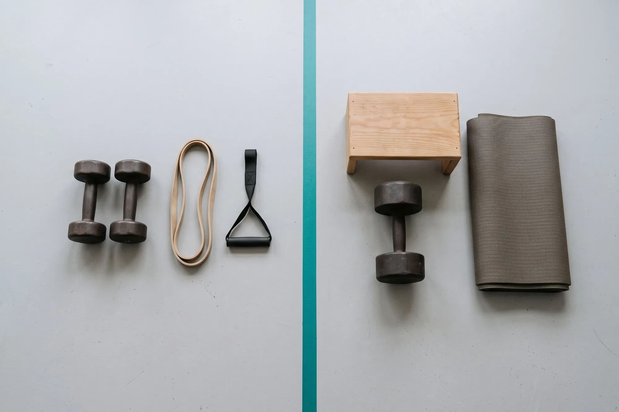 A floor split by a teal line: dumbbells, a band and a handle on one side, a heavier dumbbell, a step box and a mat on the other