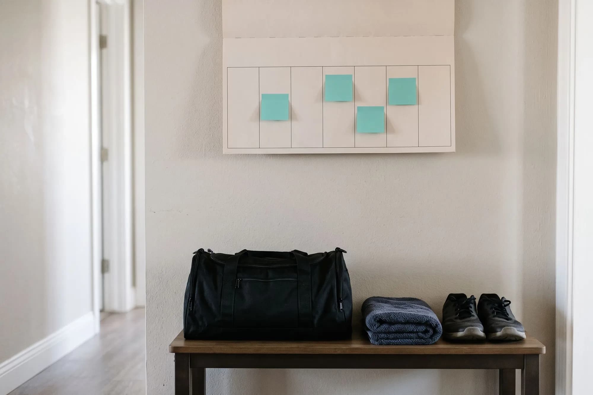 A weekly wall calendar with teal sticky notes on four days, above a gym bag, a folded towel and training shoes