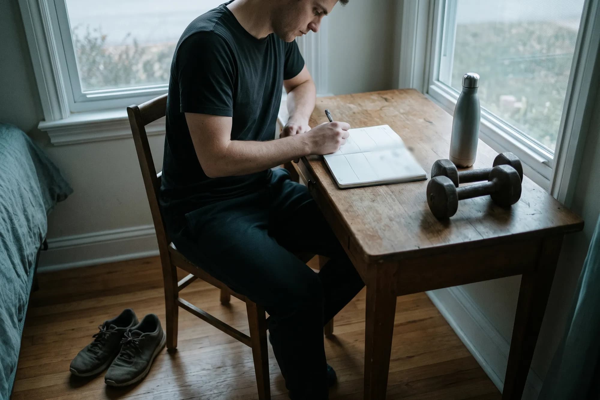 A man at a desk by a window writing his training week in a notebook, with dumbbells and a water bottle beside him